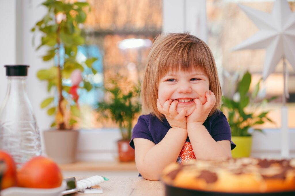 pexels-photo-1912868-1912868 A happy young girl smiling indoors surrounded by plants and a pastry, with natural light streaming in.